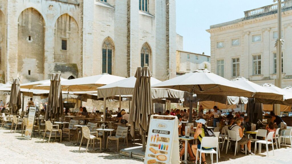 Lively summer scene at an outdoor cafe in Avignon, France, with people dining under umbrellas.